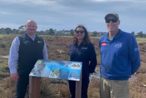 Coastal salt marsh sign with saltmarsh in background and two Country Club staff with an LLS staff member in foreground